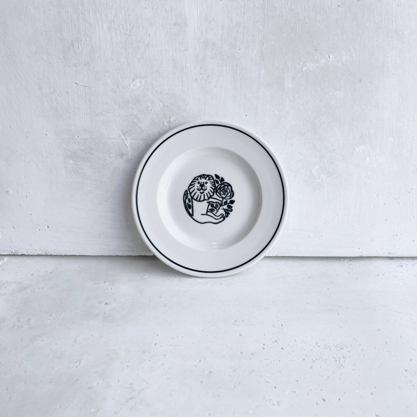 A white porcelain plate with a black line on rim and a central black illustration of a lion holding flowers, set against a textured light gray concrete background.