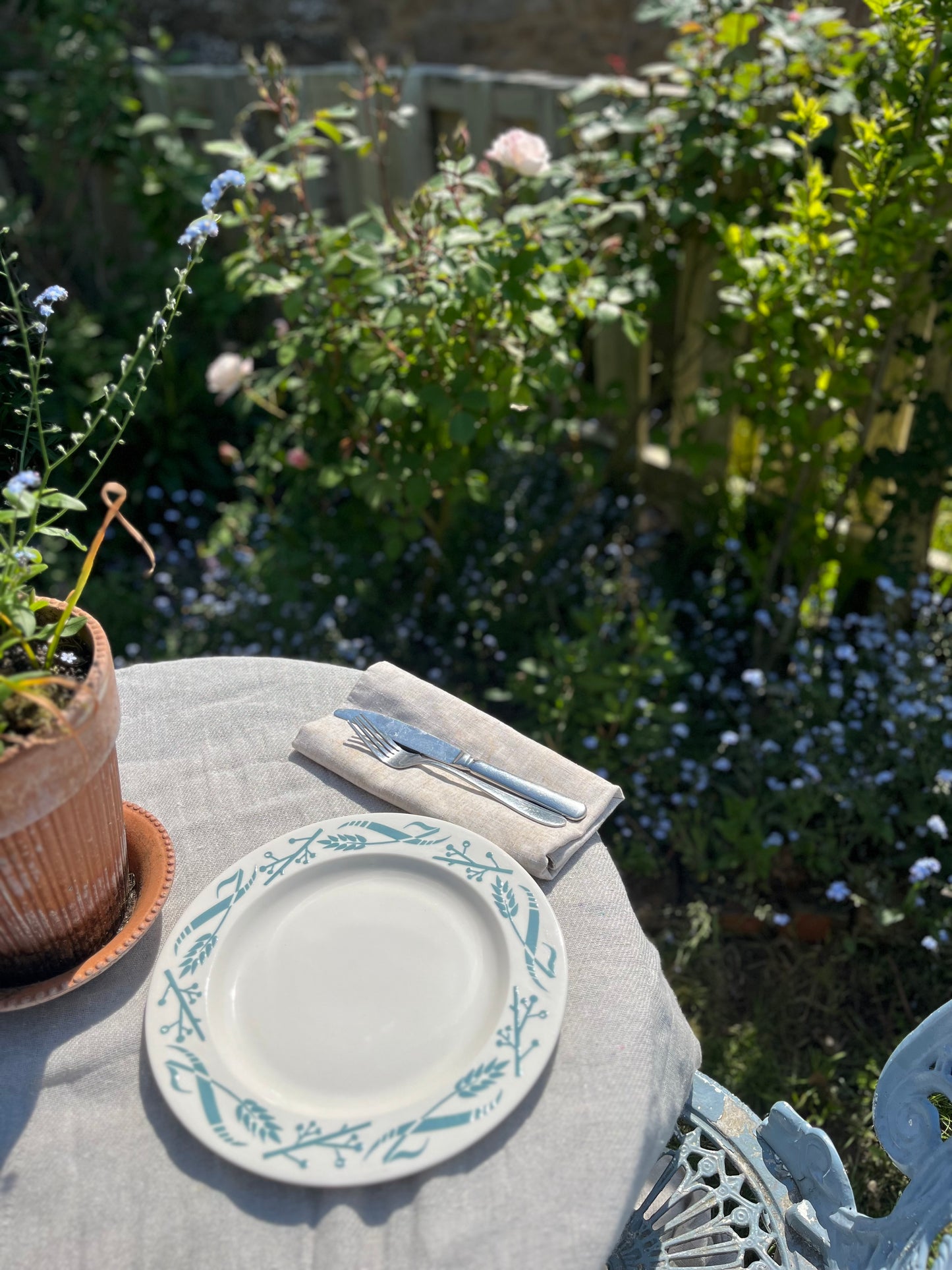 A white ceramic plate with a light blue botanical border pattern, set on a light gray tablecloth outdoors on a garden patio with a folded napkin and silver fork.