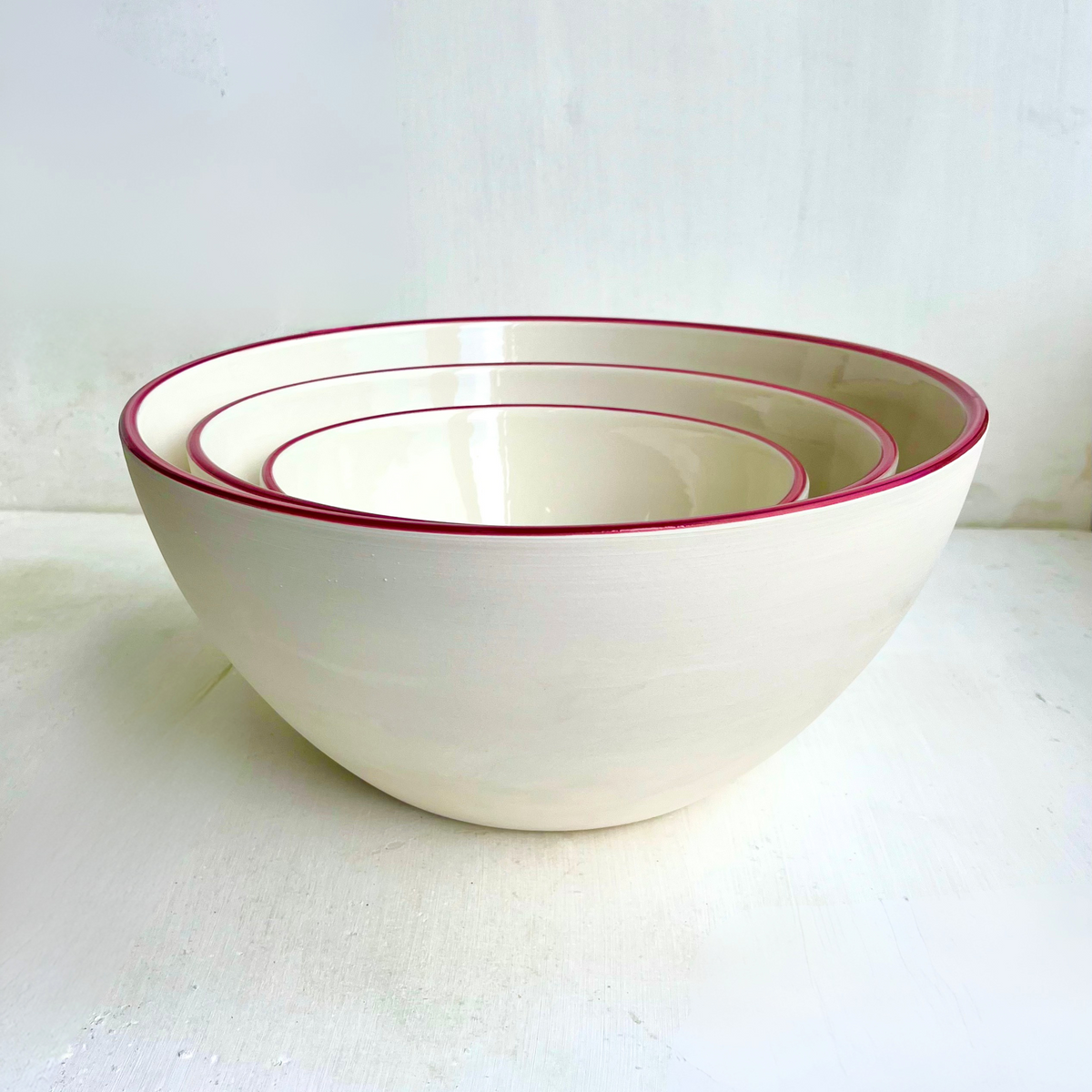Three nested ceramic bowls in a warm off-white colour, each with a thin dark burgundy red rim, set against a light, textured white background.