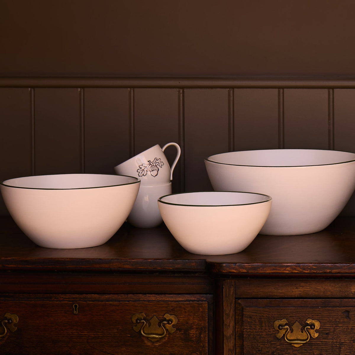 A set of three white ceramic bowls with green rim, arranged on a dark wooden chest against a dark brown paneled wall.