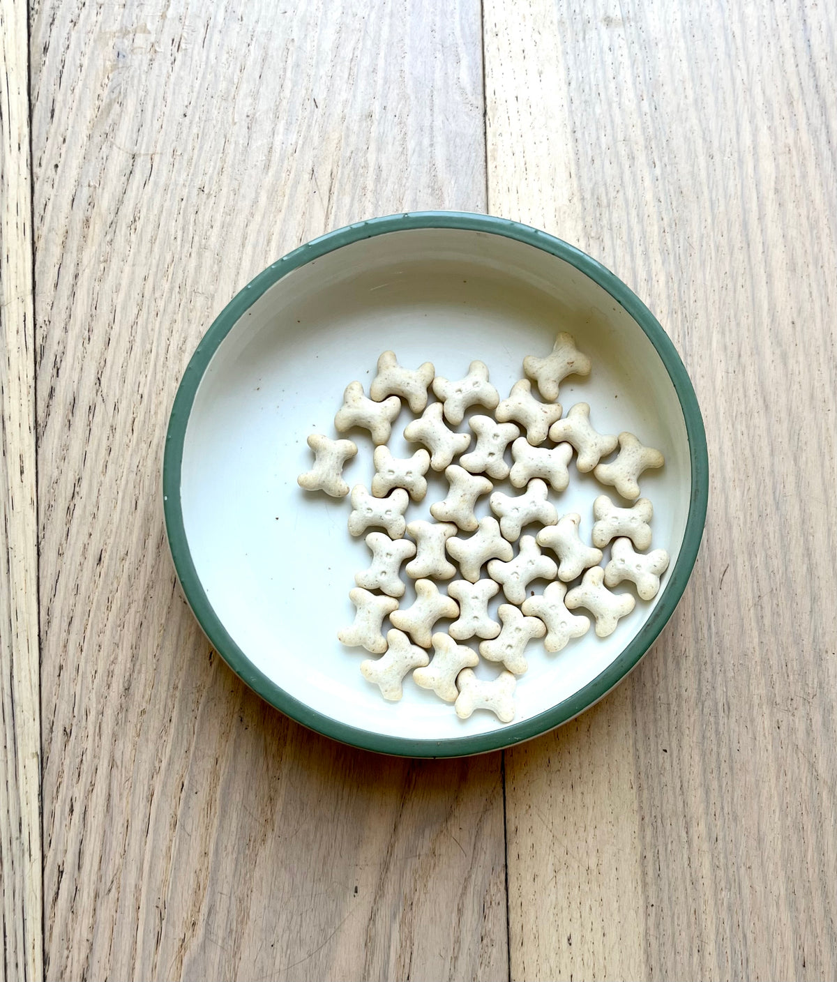 Small bone-shaped dog treats arranged in a round white bowl with a green rim, placed on a light wood tabletop.