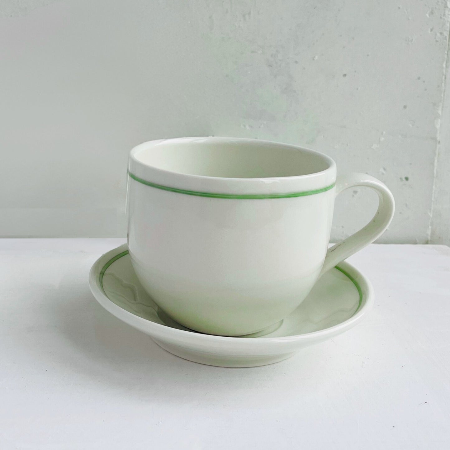 White ceramic cup with green rim and matching saucer on a white surface.